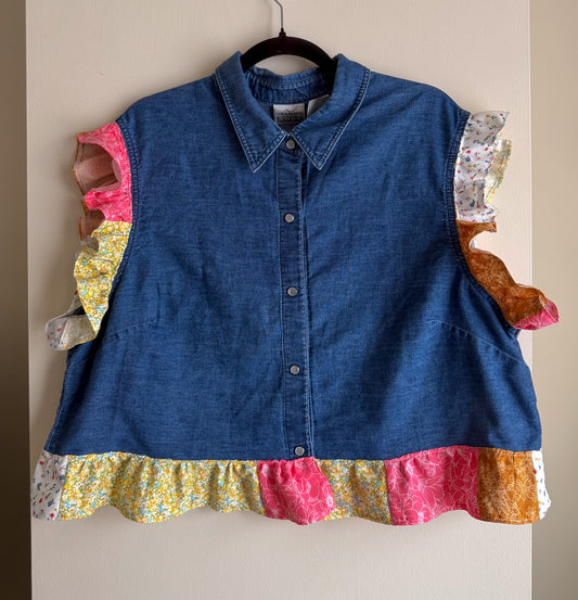 Blue denim shirt with colorful patchwork sleeves and hem on a hanger against a beige wall.