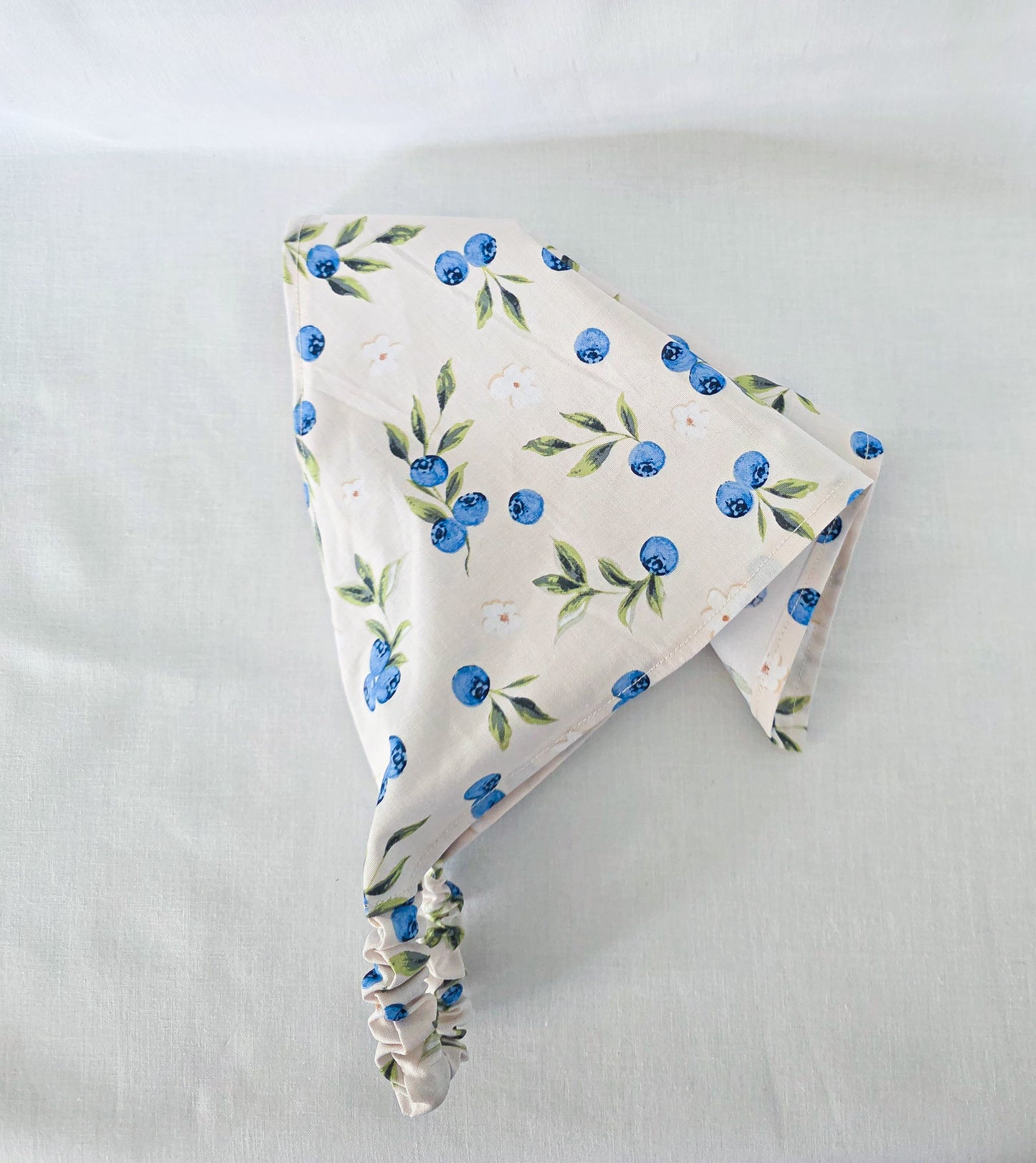 A triangular hair bandana made of cream-colored fabric with a blue and green blueberry print, featuring an elasticized strap at the bottom.