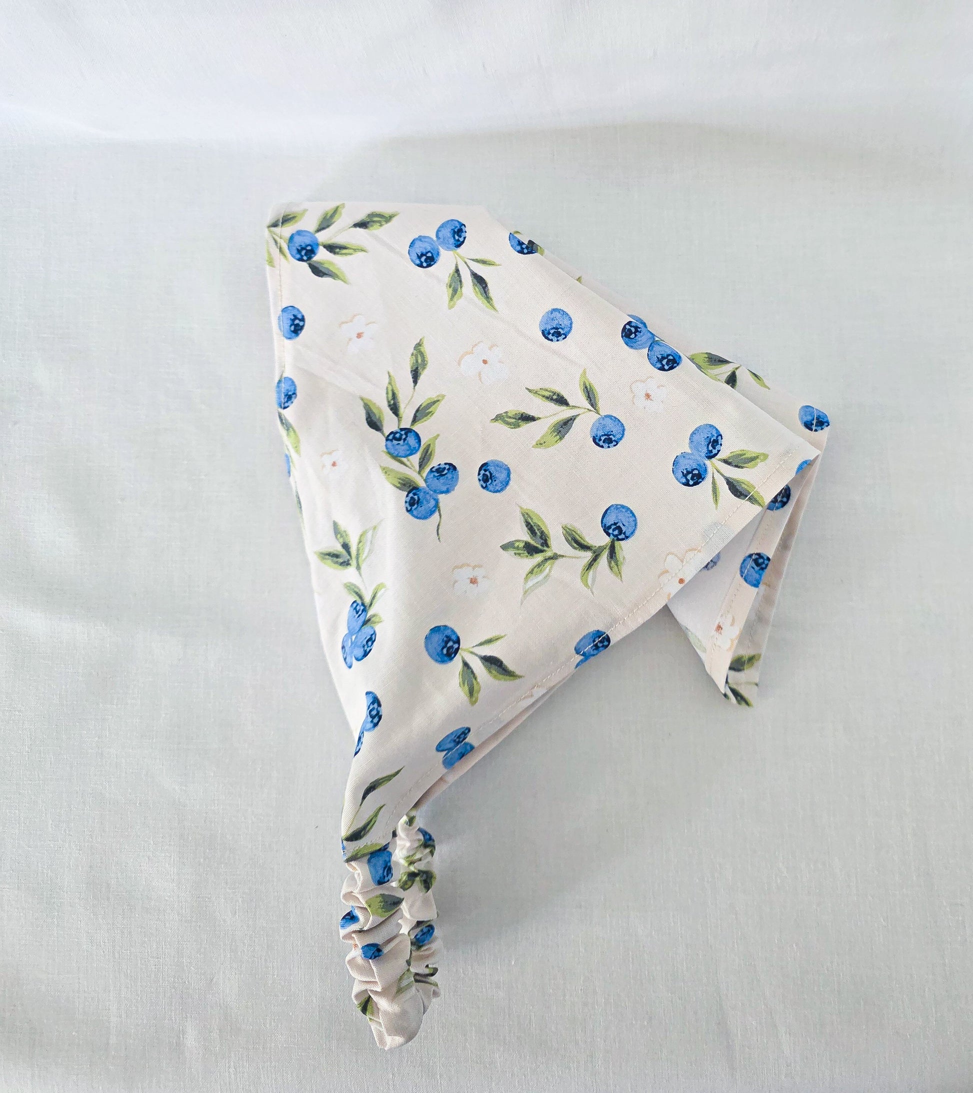A triangular hair bandana made of cream-colored fabric with a blue and green blueberry print, featuring an elasticized strap at the bottom.