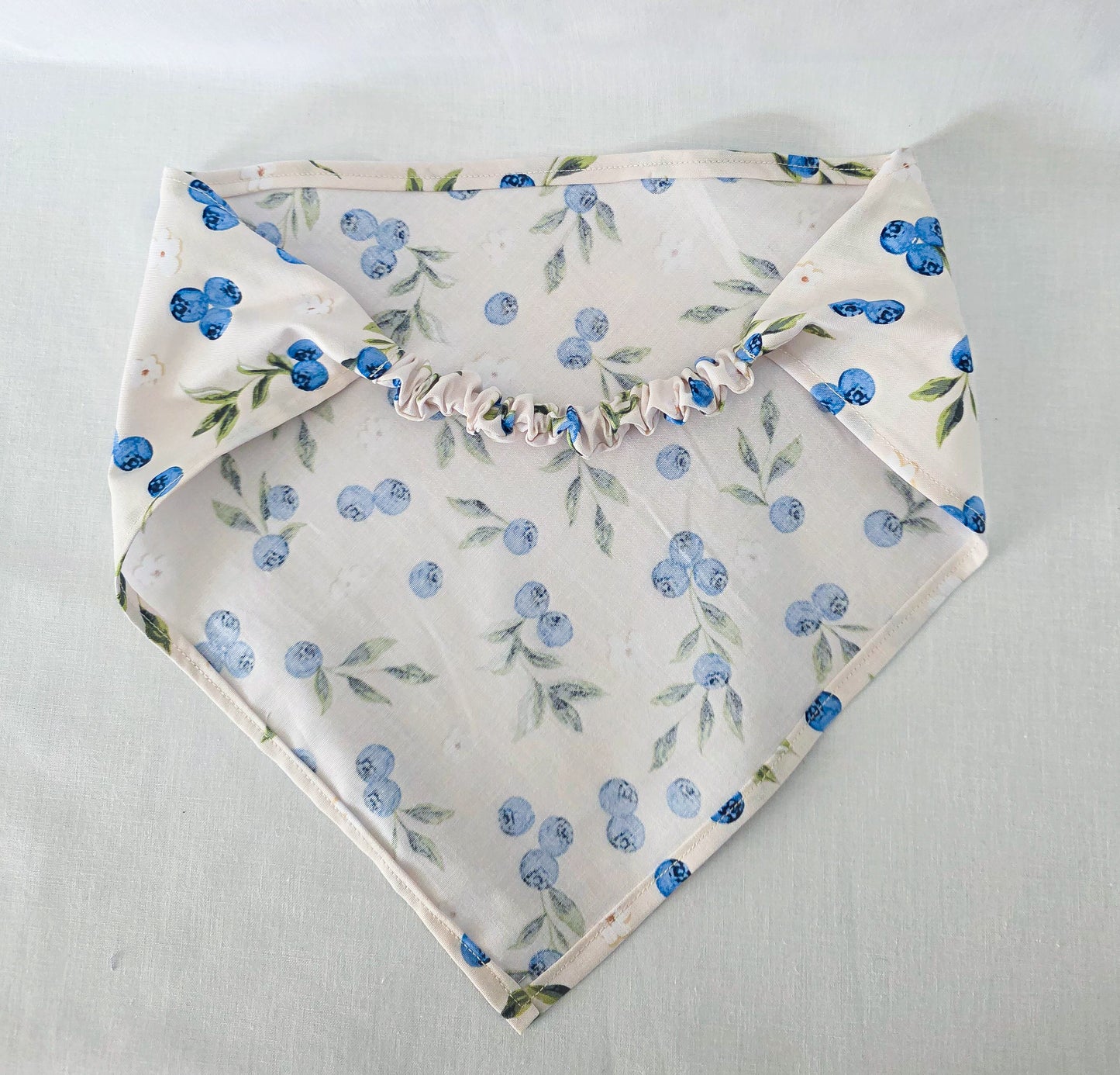 A hair bandana with a blueberry and green leaf floral print on a cream background, featuring a ruffled elastic strap, laid flat on a light fabric surface.