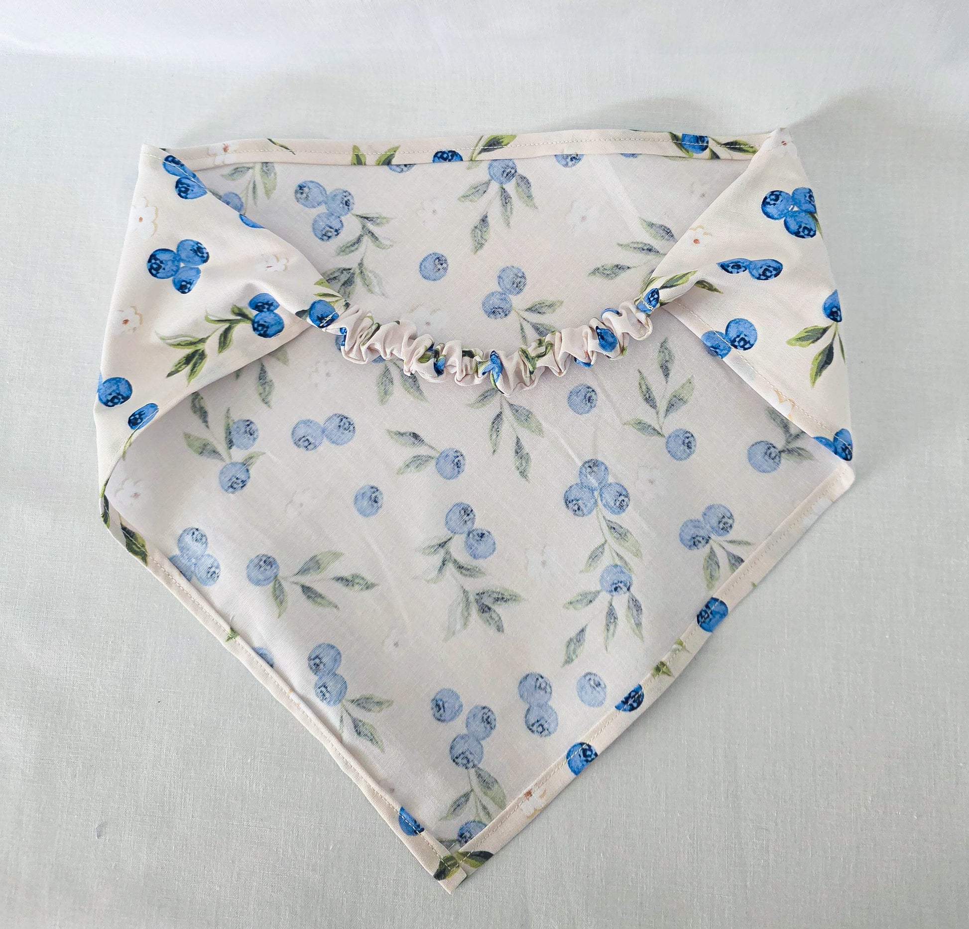 A hair bandana with a blueberry and green leaf floral print on a cream background, featuring a ruffled elastic strap, laid flat on a light fabric surface.