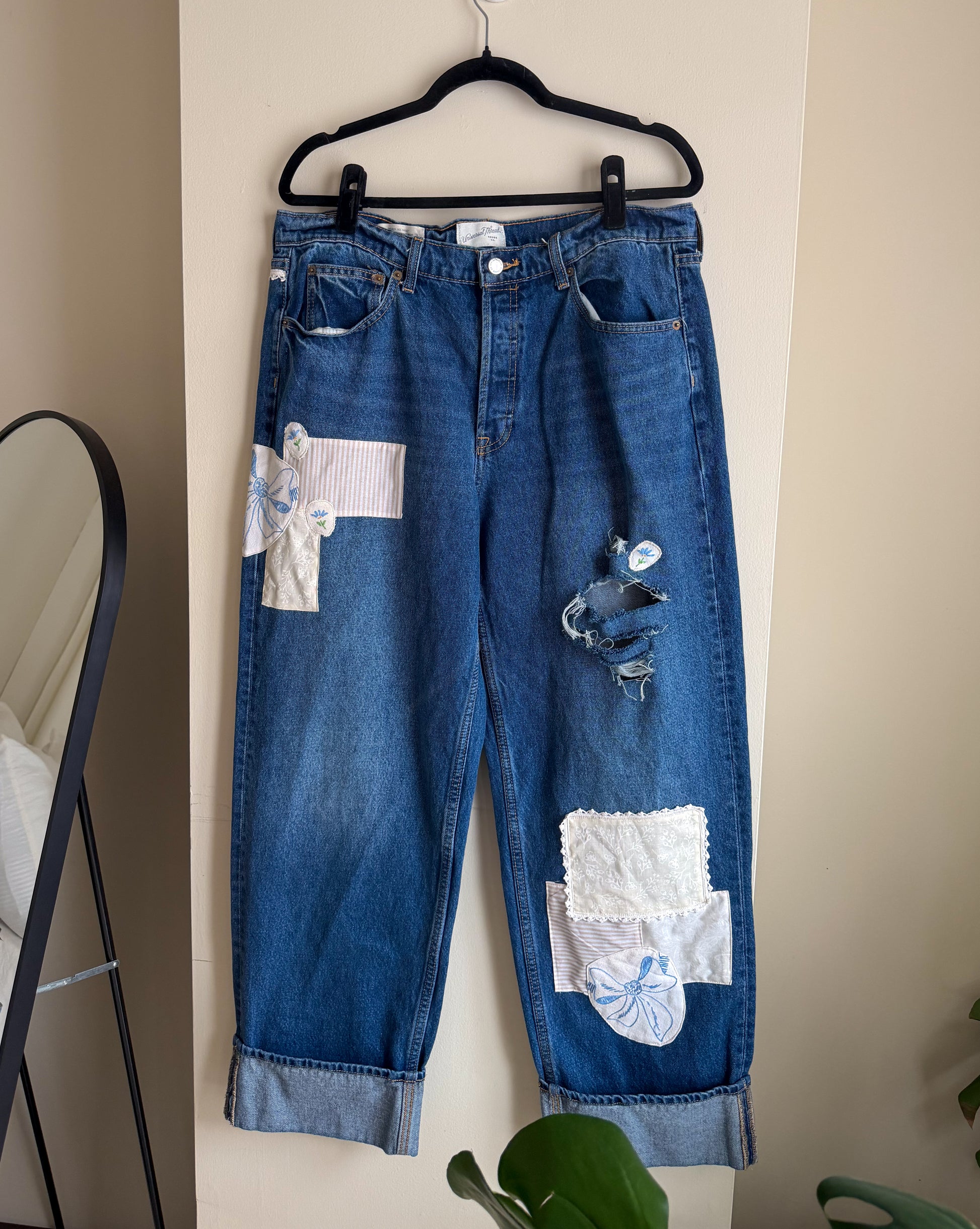 Blue jeans with patches on a hanger against a beige wall.
