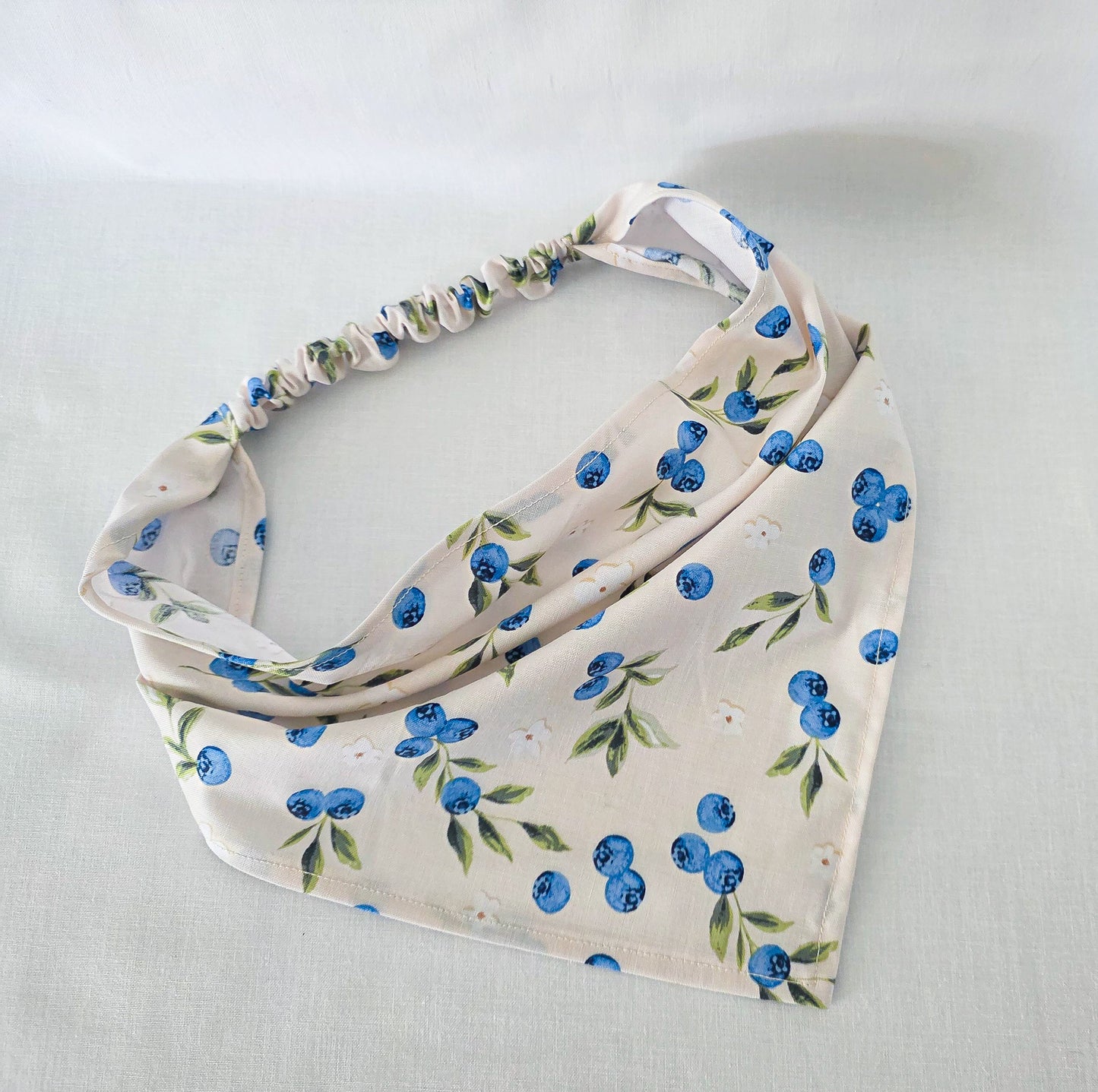 A cream-colored fabric bandana featuring a blueberry and green leaf pattern, with a gathered elastic top edge, laid flat on a light fabric background.