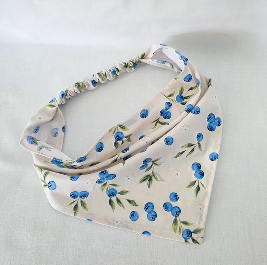 A cream-colored fabric bandana featuring a blueberry and green leaf pattern, with a gathered elastic top edge, laid flat on a light fabric background.