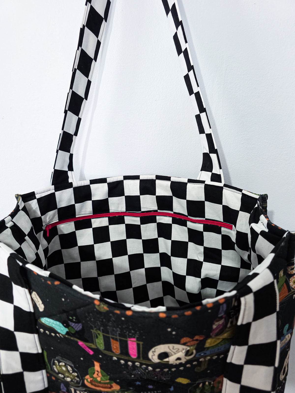 Large quilted tote bag with a witchy halloween bookshelf patterned fabric and black and white checkered straps. It is also showing the lining fabric which matches the straps and a zippered pocket.