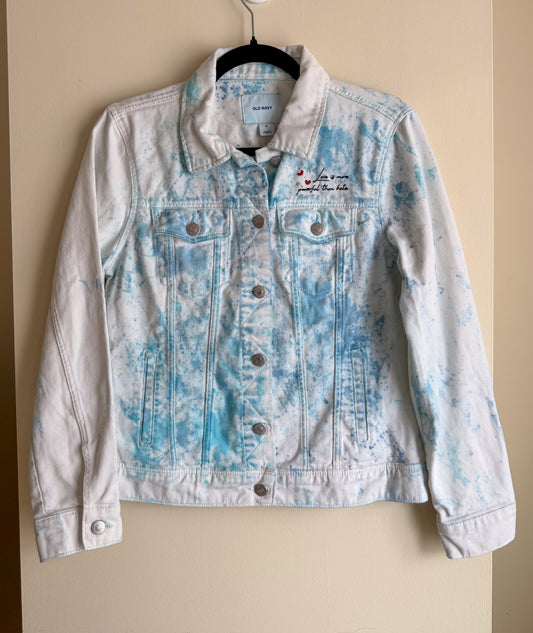 Light blue/white denim jacket with with "love is more powerful than hate" hand embroidered on the left pocket on a beige background