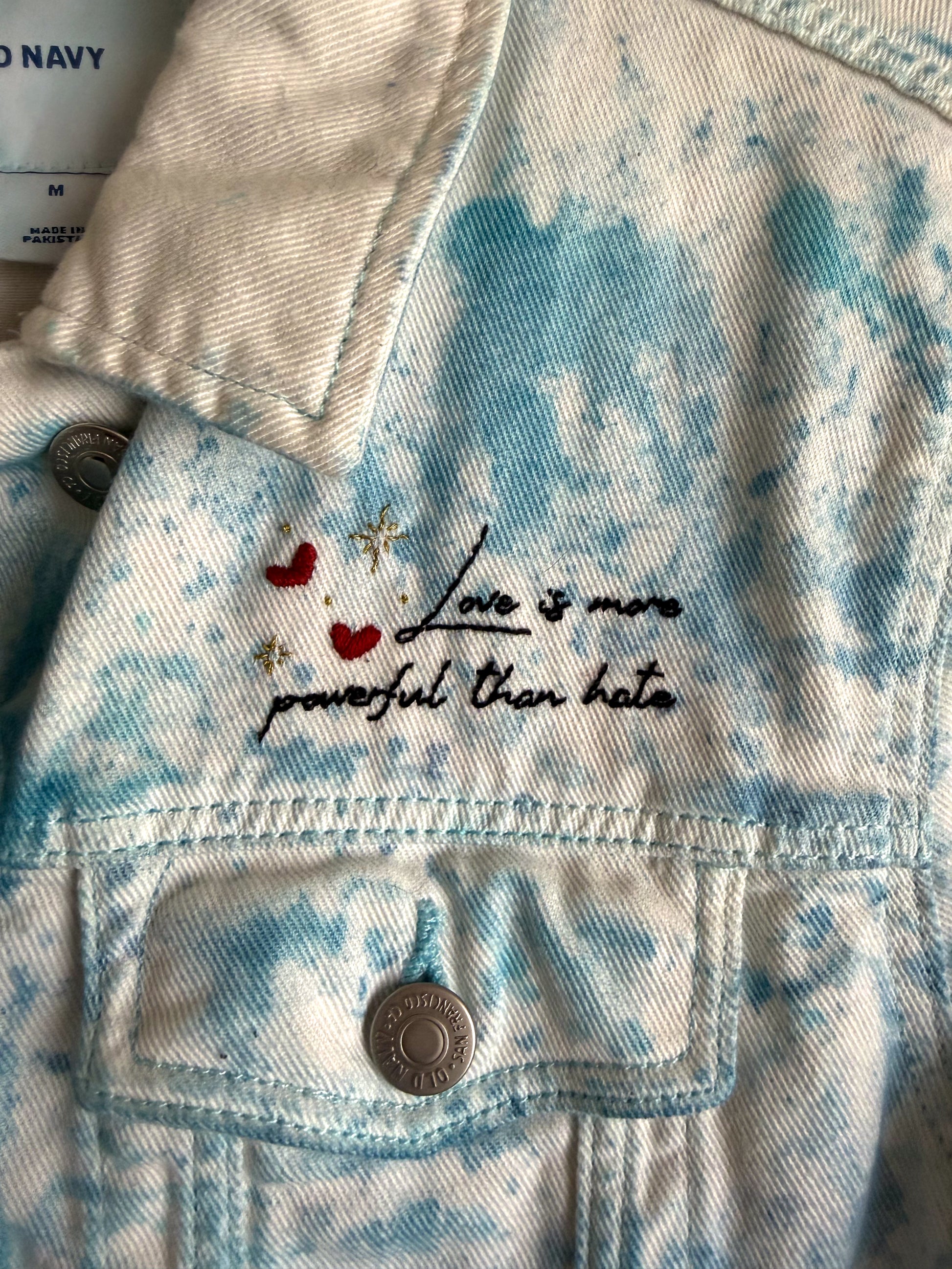 Denim jacket with embroidered text that reads "love is more powerful than hate" in cursive with hearts 