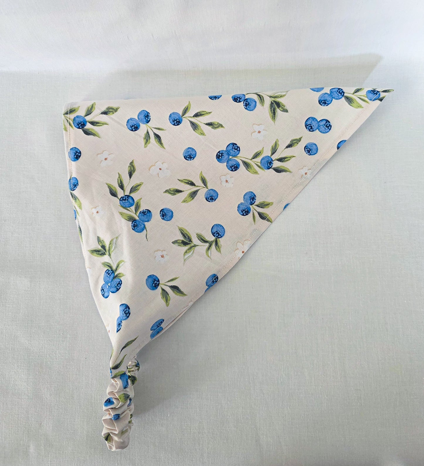 A triangular fabric hair bandana with a blueberry and green leaf pattern on a cream background.