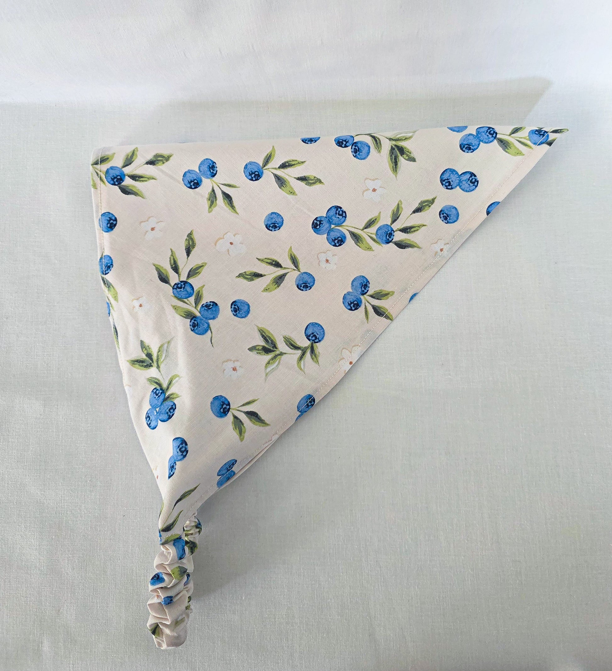 A triangular fabric hair bandana with a blueberry and green leaf pattern on a cream background.