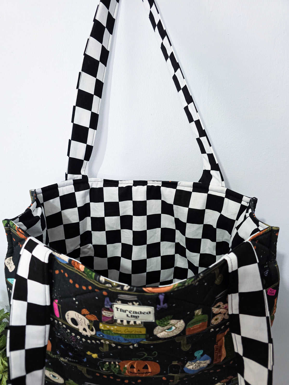 Large quilted tote bag with a witchy halloween bookshelf patterned fabric and black and white checkered straps. It is also showing the lining fabric which matches the straps